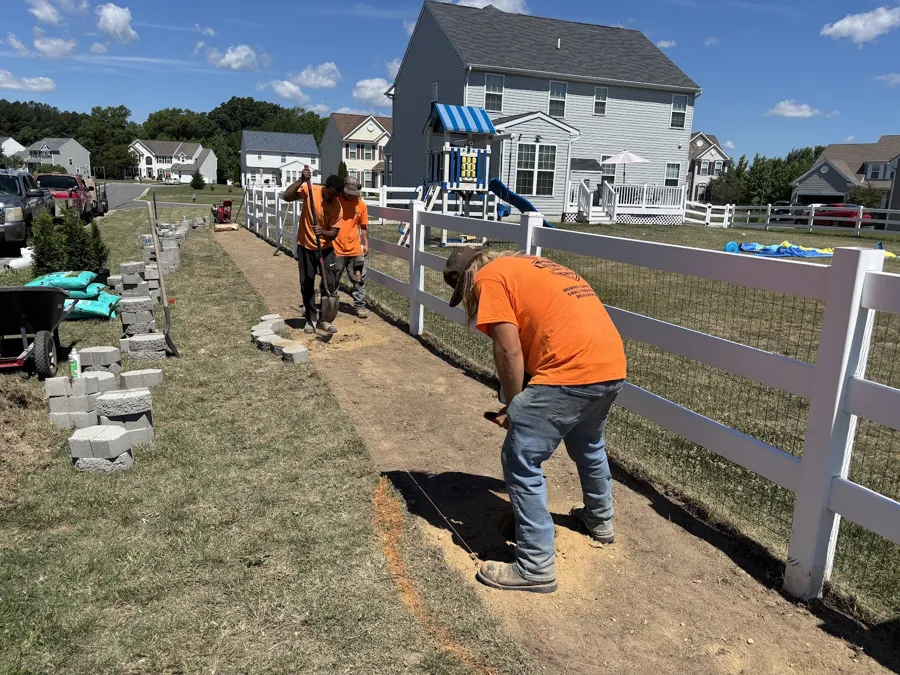 Lewis Lawn Care crew digging a level trench for block along a white vinyl fence