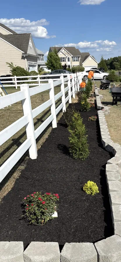 Black mulch bed with shrubs and red flowers along a white vinyl fence, edged with stacked block